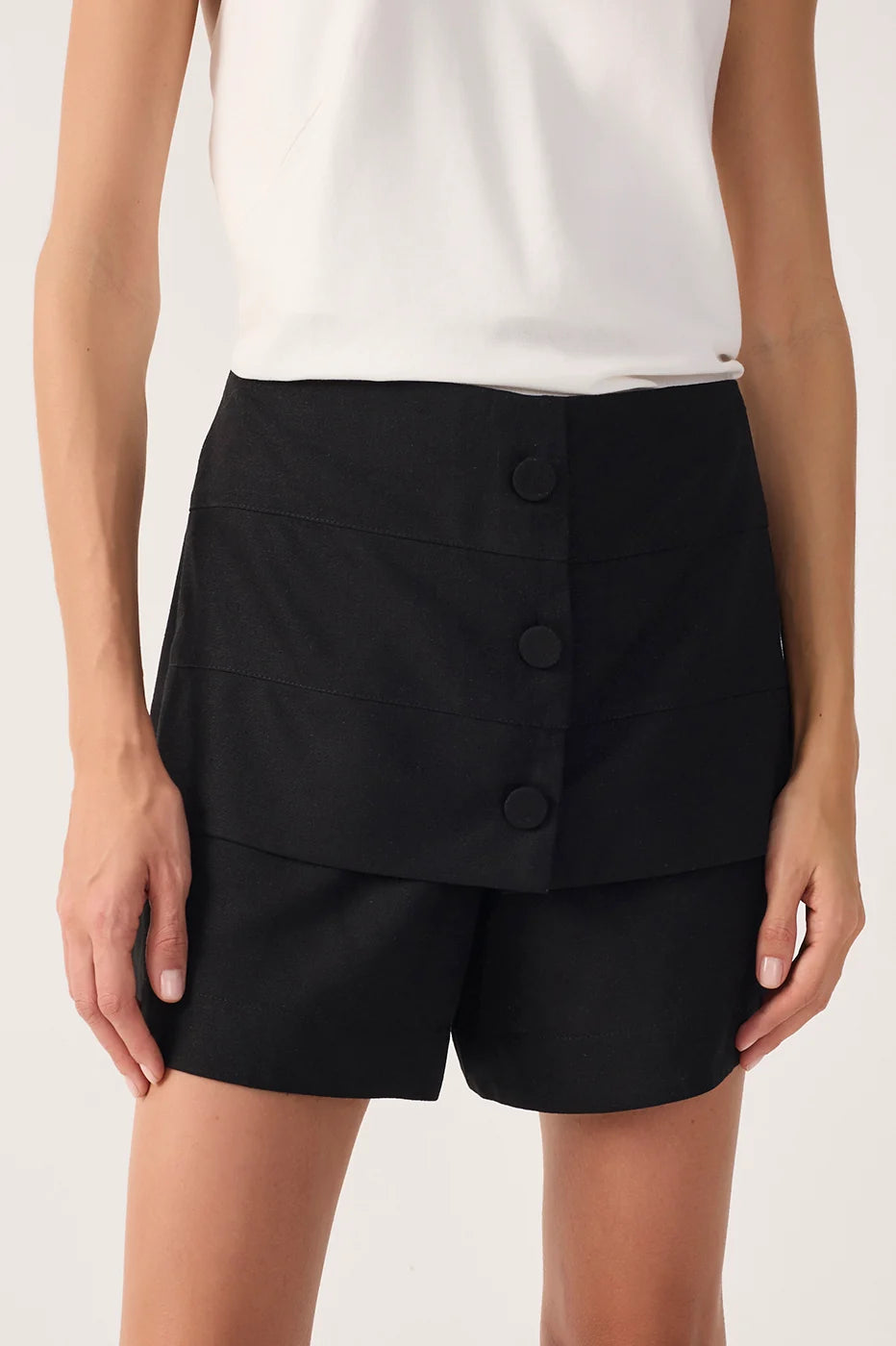 A model wearing black high-waisted shorts with a button-front wrap design and a white sleeveless top, shown against a plain light background.