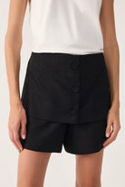 A model wearing black high-waisted shorts with a button-front wrap design and a white sleeveless top, shown against a plain light background.